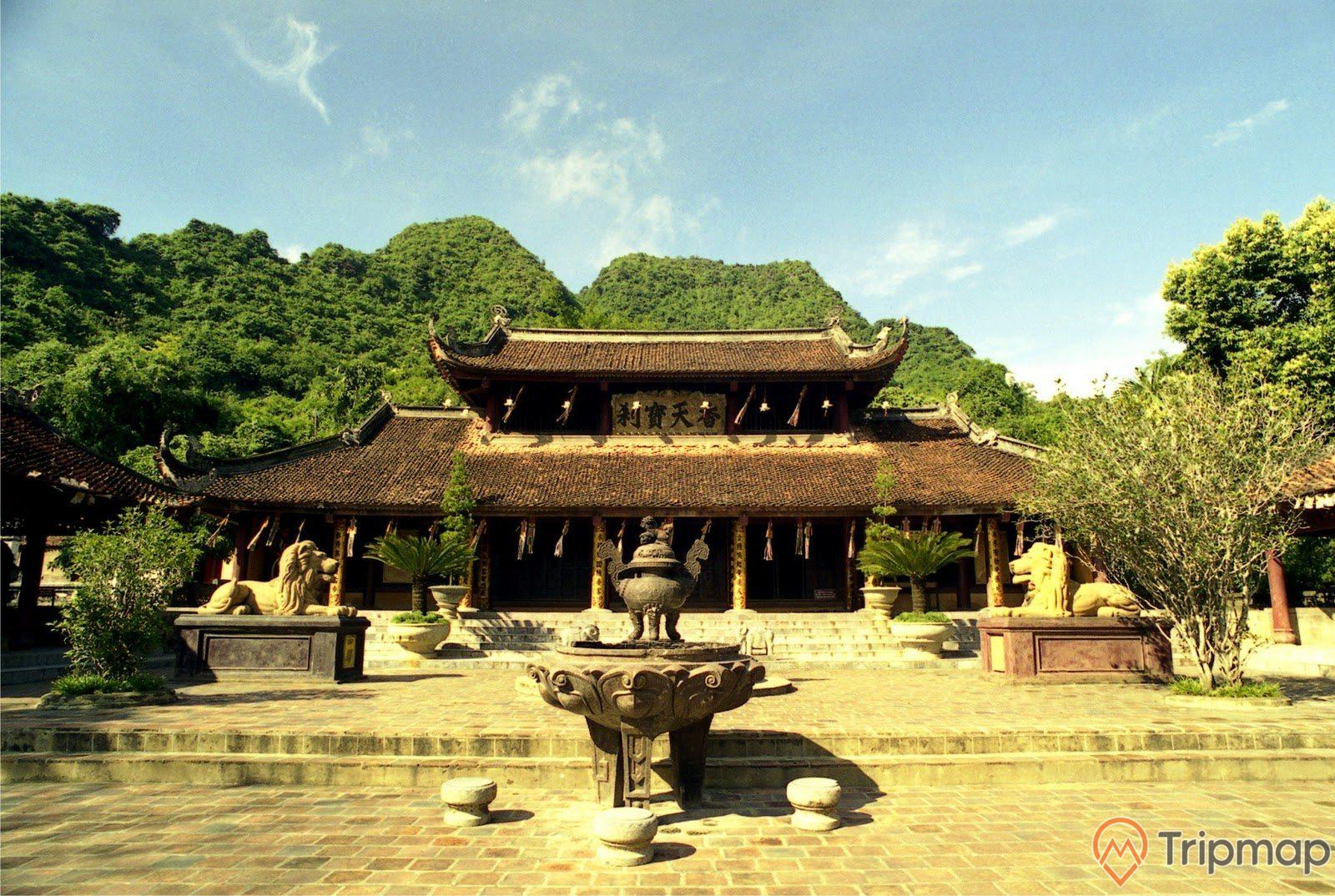 ảnh chụp chùa Thiên Trù có 2 tần mái mà ngói cũ, 3 chiếc ghế và 1 lư hương, 2 con sư tử đá màu vàng, và 6 chậu cây cảnh ở trên sân chùa, núi đồi phủ đầy cây xanh ở phía sau ngôi chùa, bầu trời trong xanh ít mây, ảnh chụp chùa Thiên Trù ngoài trời