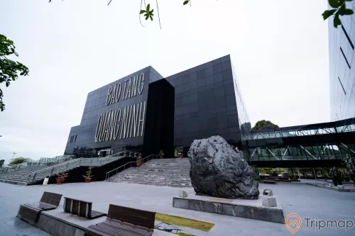 Bảo tàng Quảng Ninh, nền đường màu xám, bậc thang màu xám, nhiều chậu cây màu đỏ, nhiều ghế màu nâu, tảng đá to màu đen, nhà kính màu đen, ảnh chụp ban ngày