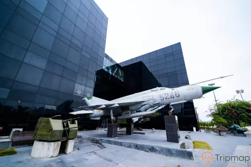 Bảo tàng Quảng Ninh, máy bay tiêm kích màu trắng, nền đường bằng gạch màu xám, tòa nhà màu đen, ảnh chụp ban ngày, cây hoa giấy