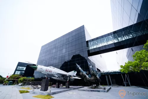 Bảo tàng Quảng Ninh, máy bay tiêm kích màu trắng, tên lửa màu xanh, nền đường bằng gạch màu xám, tòa nhà màu đen, nhà cầu, ảnh chụp ban ngày
