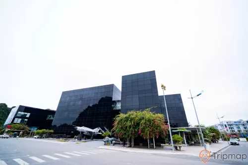 Bảo tàng Quảng Ninh, nền đường màu xám, máy bay tiêm kích màu trắng, cây hoa giấy, tòa nhà màu đen, ảnh chụp ban ngày