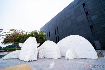 Bảo tàng Quảng Ninh, tượng cá voi màu trắng, nền gạch bằng đá màu xám, cây hoa giấy, tòa nhà màu đen, ảnh chụp ban ngày