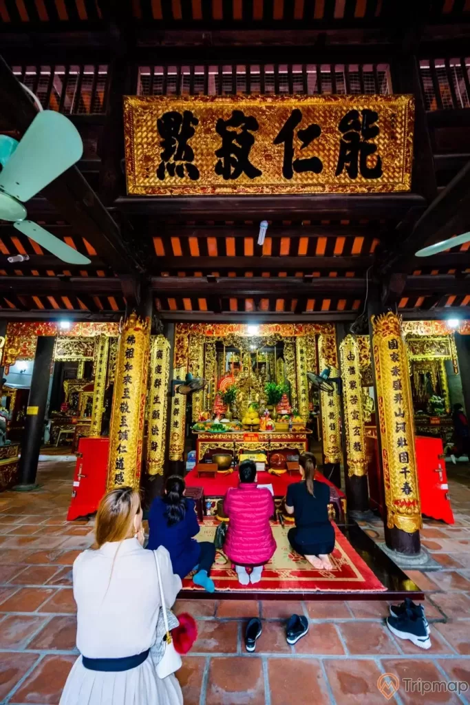 Chùa Long Tiên, nền gạch màu đỏ, nhiều người đang cầu nguyện, tượng Đức thánh Trần Hưng Đạo, nhiều cây cột nhà màu vàng có hoa văn nhiều chữ hán, cây quạt trần, trần nhà bằng gỗ màu nâu, ảnh chụp ban ngày