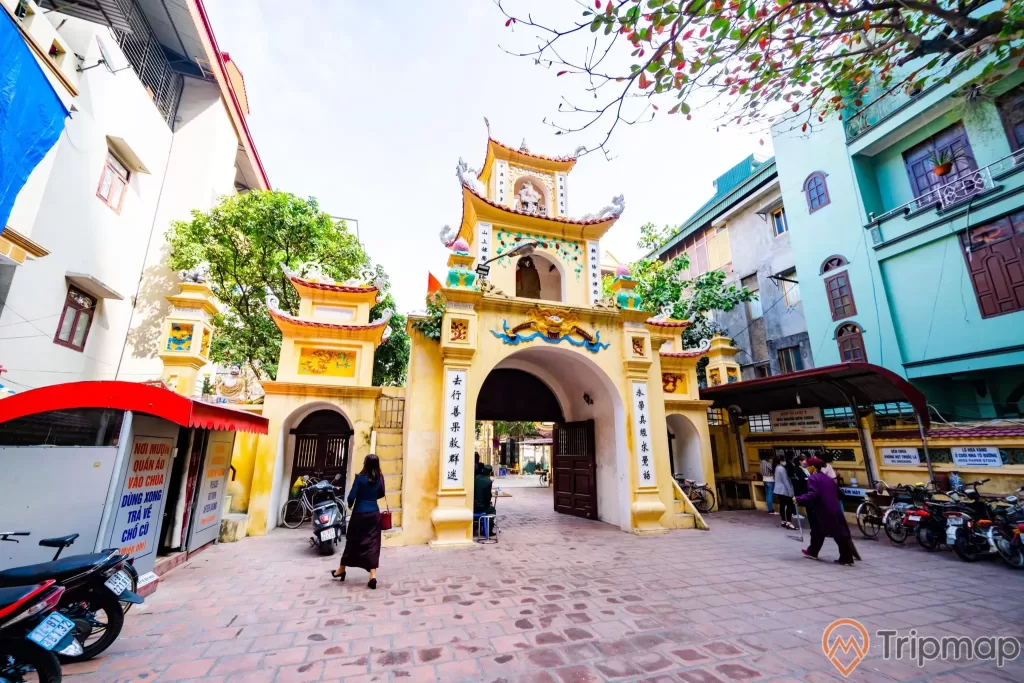 Chùa Long Tiên, cổng Tam Quan Nội, nền gạch màu đỏ, cổng sơn màu vàng, nhiều xe mày, nhà dân sơn màu xanh, ảnh chụp ban ngày