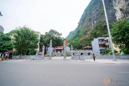 Đền thờ Đức Ông Trần Quốc Nghiễn, nền đường màu xám, cây cột đèn, lá cờ Việt Nam, nhiều cây xanh, ngọn núi đá có cây xanh, ảnh chụp ban ngày