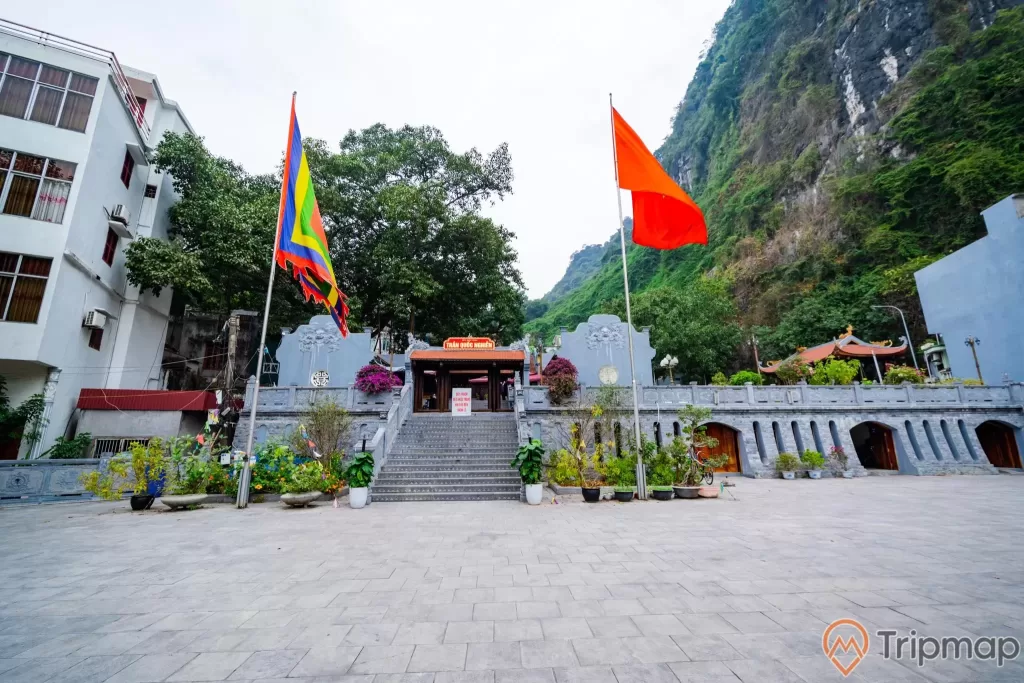 Đền thờ Đức Ông Trần Quốc Nghiễn, lá cờ Việt Nam, nền đường bằng gạch màu xám, bậc thang màu xám, nhiều chậu cây, ngọn núi đá có cây xanh, ảnh chụp ban ngày