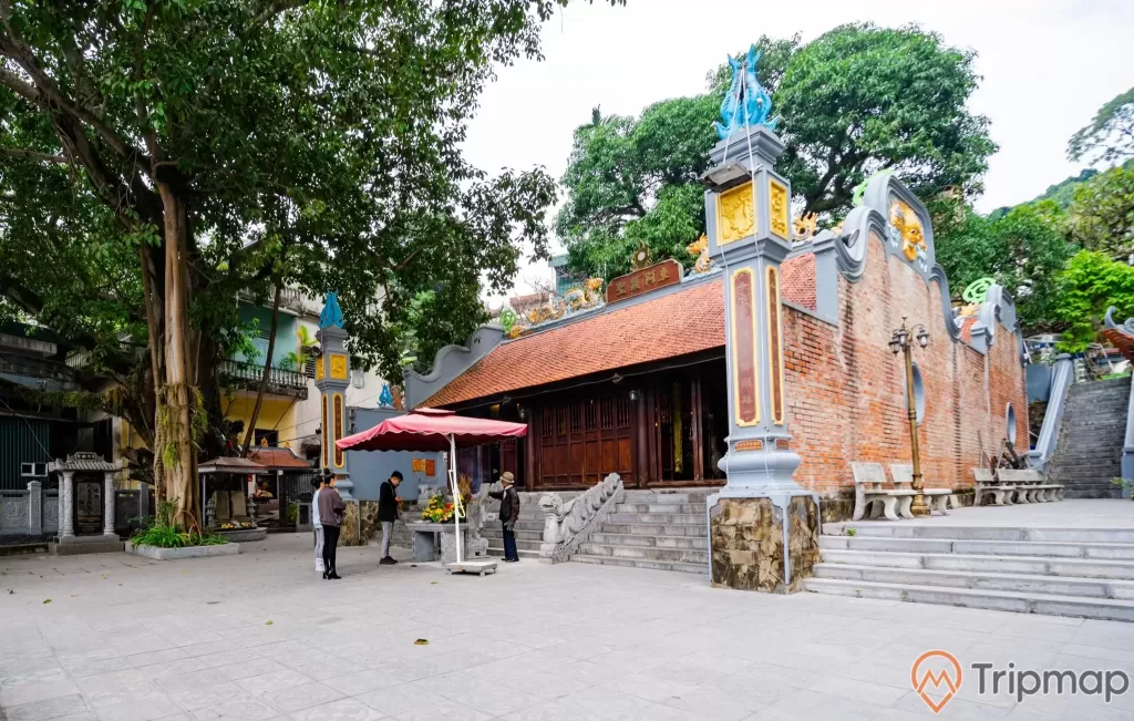 Đền thờ Đức Ông Trần Quốc Nghiễn, nền đường màu xám, bậc thang màu xám, đền thờ bằng gạch màu đỏ, nhiều cây xanh, ảnh chụp ban ngày