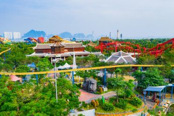 Khu vui chơi giải trí Sun World Ha Long Complex, nhiều cây xanh, tòa nhà có mái ngoái màu nâu, tảng đá hình mặt người, nhiều ngọn núi đá có cây xanh phía xa, trời xanh nhiều mây, ảnh chụp ban ngày, ảnh chụp từ trên cao