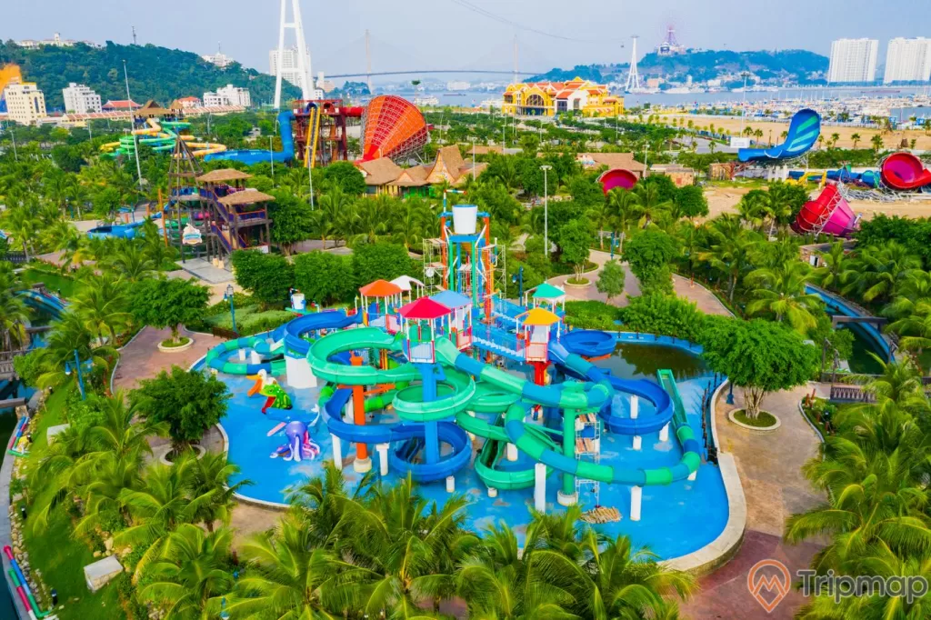 Khu vui chơi giải trí Sun World Ha Long Complex, ống trượt màu xanh trên bể bơi, nhiều cây xanh, nền gạch màu đỏ, cầu bãi cháy ở phía xa, ảnh chụp ban ngày, ảnh chụp từ trên cao