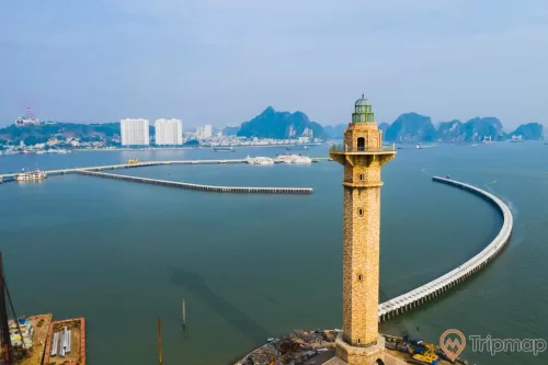 Ngọn hải đăng Sun World, ngọn hải đăng màu vàng, cầu tầu màu trắng , mặt nước biển màu xanh, nhà cao tầng màu trắng ở phía xa, nhiều ngọn núi đá có cây xanh phía xa, trời xanh nhiều mây, trời nắng, ảnh chụp ban ngày