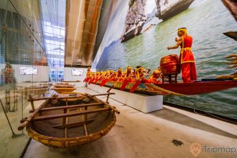 Bảo tàng Quảng Ninh, biển cả và tự nhiên, thuyền màu nâu, bức trangh hòn trống mái, nhiều người đang chèo thuyền màu đỏ, nhiều người mặc áo đỏ, người đàn ông mặc áo đỏ đánh trống, nền nhà màu nâu nhạt