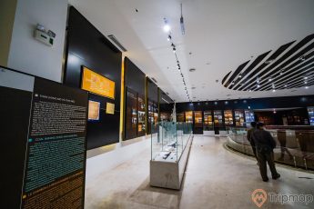 Khu tái hiện lịch sử hình thành mỏ than, bảo tàng Quảng Ninh, nhiều tủ kính trưng bày hiện vật, người đang đi trên nền gạch có hoa văn, bảng giới thiệu màu đen, trần nhà màu trắng