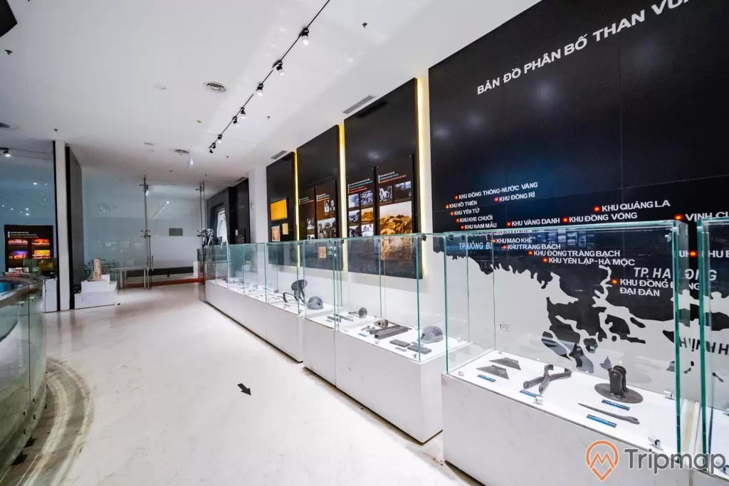 Khu tái hiện lịch sử hình thành mỏ than, bảo tàng Quảng Ninh, nhiều dụng cụ được bày trong tủ kính, bản đồ màu đen, nền gạch màu trắng có mũi tên màu đen nằm trên, trần nhà màu trắng