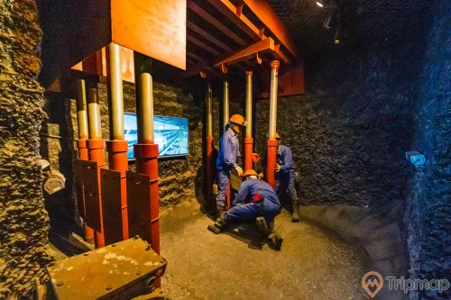Khu tái hiện lịch sử hình thành mỏ than, bảo tàng Quảng Ninh, mô hình nhiều người công nhân mặc quần áo xanh đang làm việc, nền đất màu nâu, ánh sáng vàng, màn hình ti vi