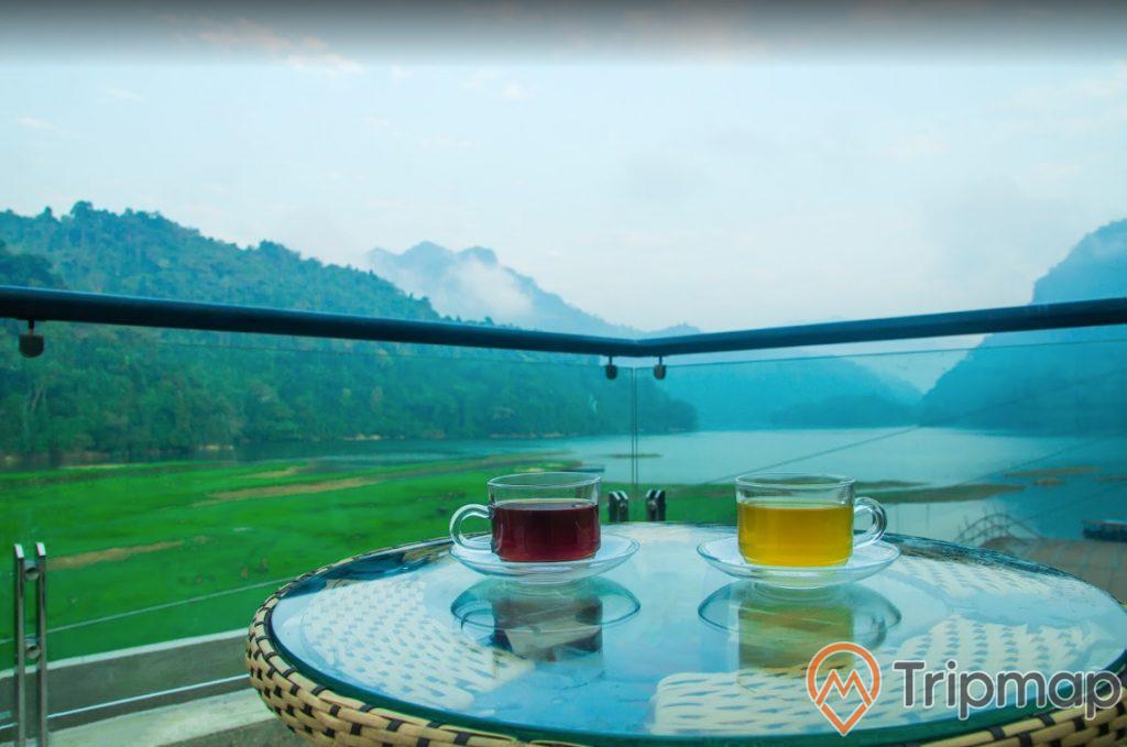 view ngắm nhìn cảnh tại ao tiên, 2 cốc trà trên mặt bằng kính, đồi núi cây cối ở phía xa, bầu trời nhiều mây, ảnh chụp ngoài trời