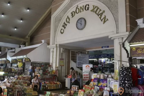 chiếc đồng hồ và dòng chữ chợ đồng xuân ở phía trên cửa ra vào chợ, 2 người phụ nữ đang đi và 1 người đàn ông đang ngồi cạnh quán tạp hoá