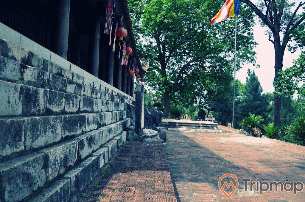 khuôn viên sân chùa Trăm Gian, lá cờ nhiều màu sắc đang bay phất phới, lồng đèn treo dưới mái hiên chùa, cây cối xanh tươi trước sân chùa