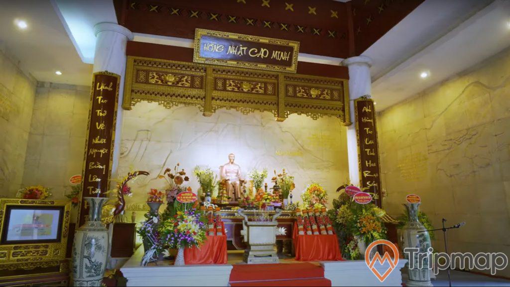 điện thờ chủ tịch Hồ Chí Minh tại di tích Pác Bó, ảnh chụp trong nhà, tượng bà Hồ đang ngồi trên ghế, lư hương ở giữa và giỏ hoa trên điện thờ, 2 chiếc bình bằng gốm xứ, ánh đèn vàng trong nhà