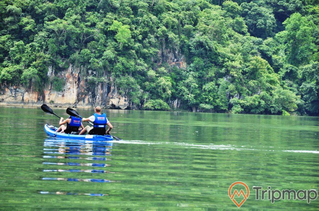 du khách tham quan tại hồ Ba Bể, 2 du khách mặc áo phao đang ngồi chèo trên thuyền màu xanh, cây cối xanh tươi trên vách núi đá, ảnh chụp ngoài trời
