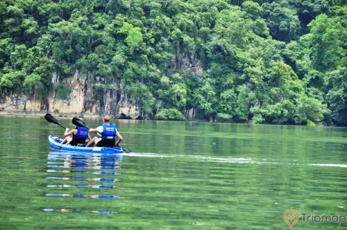 du khách tham quan tại hồ Ba Bể, 2 du khách mặc áo phao đang ngồi chèo trên thuyền màu xanh, cây cối xanh tươi trên vách núi đá, ảnh chụp ngoài trời
