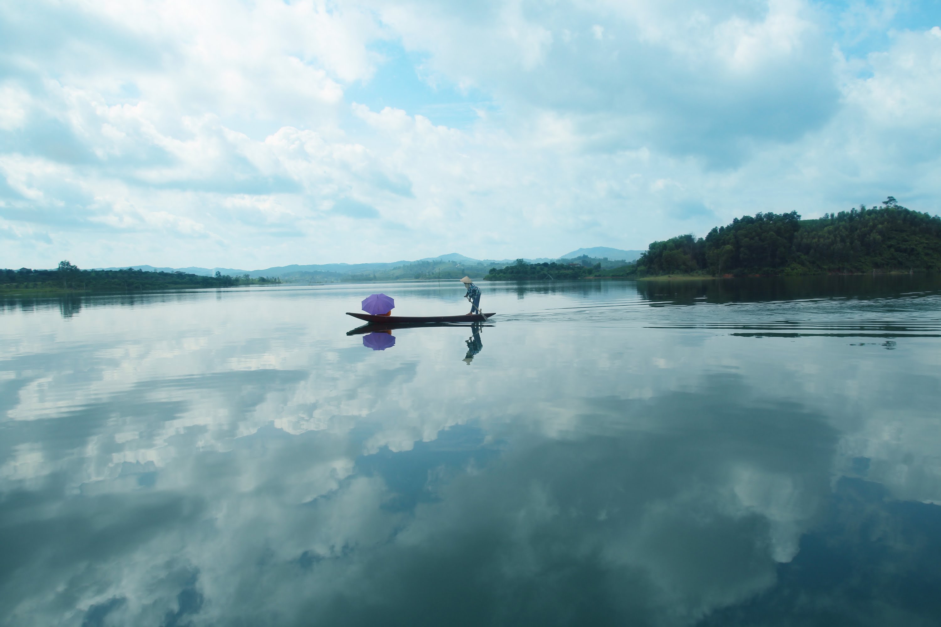 người chèo thuyền giữa Hồ Cấm Sơn, một người đang đứng trên thuyền chèo lái trên mặt hồ, núi đồi ở phía xa, bầu trời nhiều mây, ảnh chụp ngoài trời
