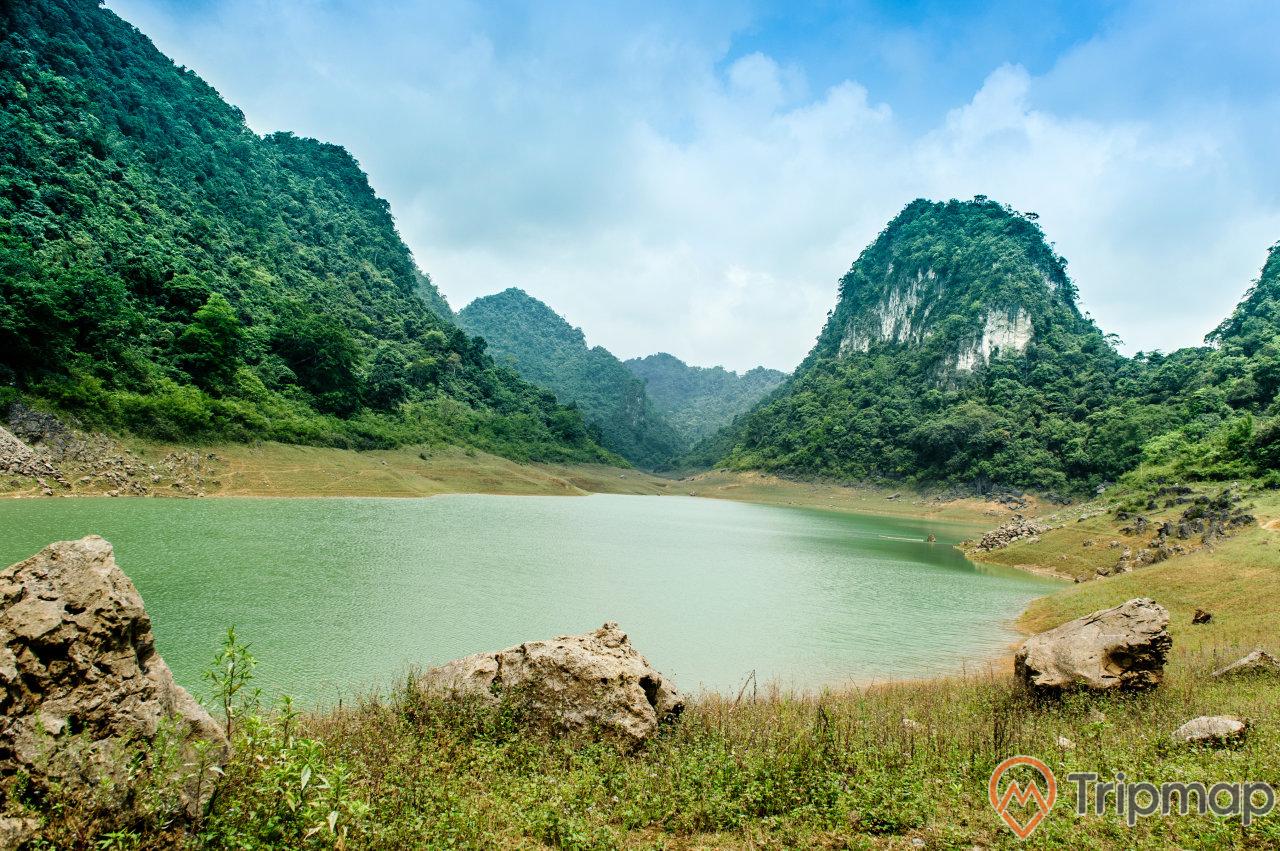 Khung cảnh Hồ Thang Hen, cây cỏ và tảng đá bên bờ hồ, đồi núi cây xanh ở phía xa, bầu trời nhiều mây, ảnh chụp ngoài trời