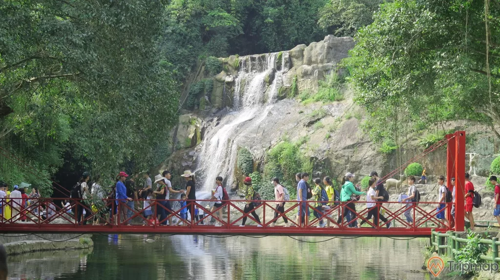 Cây cầu bắc ngang qua hồ khu du lịch Ao Vua, mội người đang đi trên cầu màu đỏ, thác nước đang chảy xuống hồ nước, cây cối mọc trên vách núi đá, ảnh chụp ngoài trời