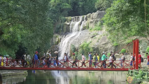 Cây cầu bắc ngang qua hồ khu du lịch Ao Vua, mội người đang đi trên cầu màu đỏ, thác nước đang chảy xuống hồ nước, cây cối mọc trên vách núi đá, ảnh chụp ngoài trời
