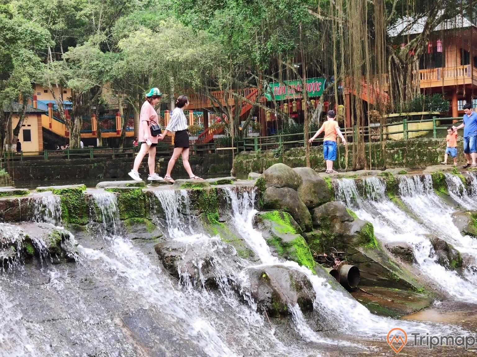 du khách tham quan hồ nước khu du lịch ao vua, 5 người đang đi trên bờ hồ có thác nước đang chảy, cây cối và nhà cửa gần hồ nước