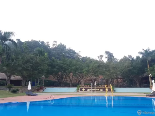 Bể bơi trong khu du lịch Thác Đa, bể bơi màu xanh, có 2 bức tượng màu vàng, chậu cây cạnh bậc thềm cây cối mọc quanh bể bơi, bầu trời nhiều mây,