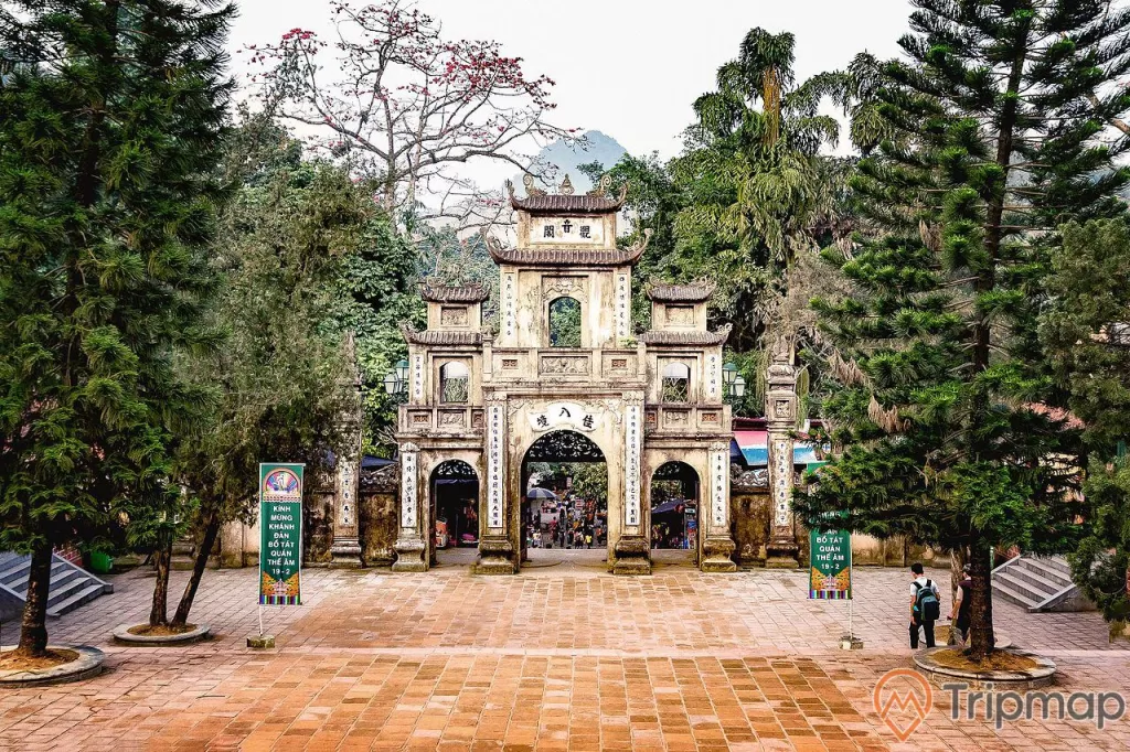 ảnh chụp khuôn viên và cổng chùa Hương, 2 người đang đi bộ, 2 cây tùng, 2 tấm biển poster, cây cối phía bên ngoài chùa Hương, ảnh chụp ngoài trời