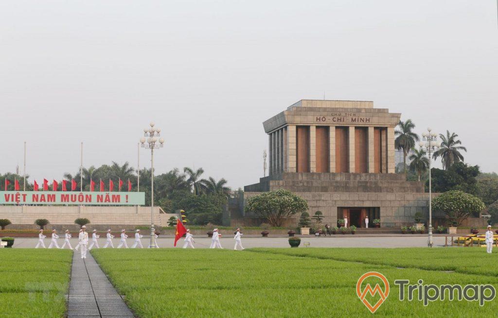 Lính diễu hành lăng bác cầm cờ đỏ sang vàng Việt Nam ở trong quảng trường lăng bác, dòng chữ "Việt Nam muôn năm", 2 người lính gác 2 cây đèn sân cỏ trước trước quảng trường lăng bác, cây cối cạnh lăng bác mọc cao xanh um tùm
