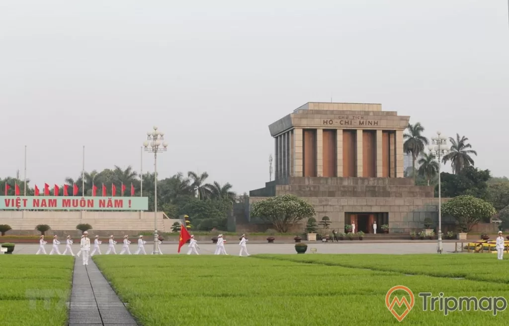 Lính diễu hành lăng bác cầm cờ đỏ sang vàng Việt Nam ở trong quảng trường lăng bác, dòng chữ "Việt Nam muôn năm", 2 người lính gác 2 cây đèn sân cỏ trước trước quảng trường lăng bác, cây cối cạnh lăng bác mọc cao xanh um tùm