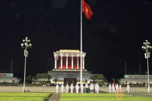 cột cờ Việt Nam bay phất phới, 2 cây cột lớn, bãi cỏ xanh, những người lính áo trắng đang tham dự lễ hạ cờ, bầu trời tối tại quảng trường lăng bác