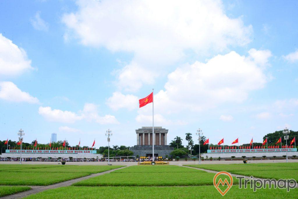 14 lá cờ Việt Nam ở phía xa gần Lăng chủ tịch Hồ Chí Minh, 1 lá cờ lá cờ đỏ sao vàng đang bay phất phới, 4 cây đèn lớn và sân ở cỏ ở trước lăng bác, cây cối mọc um tùm quanh Lăng Bác, bầu trời trong xanh có mây
