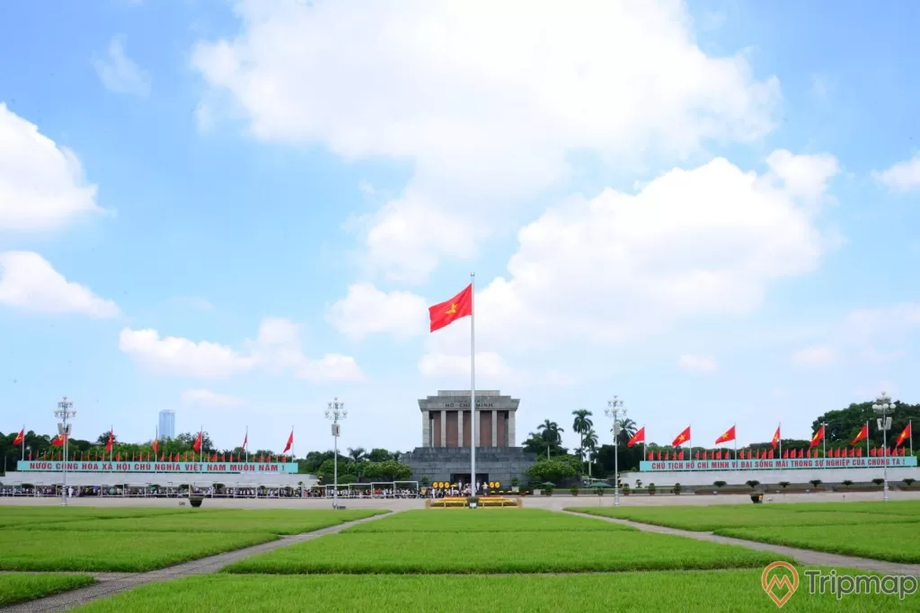 14 lá cờ Việt Nam ở phía xa gần Lăng chủ tịch Hồ Chí Minh, 1 lá cờ lá cờ đỏ sao vàng đang bay phất phới, 4 cây đèn lớn và sân ở cỏ ở trước lăng bác, cây cối mọc um tùm quanh Lăng Bác, bầu trời trong xanh có mây