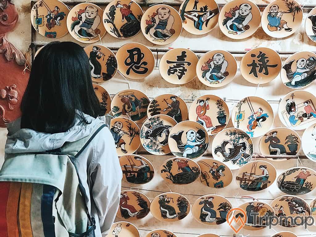 cô gái đang đứng nhìn những cái đĩa gốm sứ hình vẽ tranh đông hồ treo trên tường