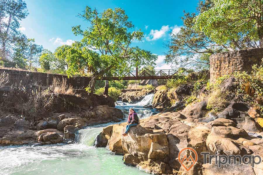 quang cảnh dòng nước tại Thác Ba Tia, bầu trời trong xanh ít mây, cây cối xanh tươi bên bờ suối, cô gái ngồi trên phiến đá cạnh dòng suối, ảnh chụp ngoài trời