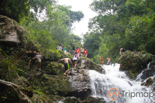 du khách trải nghiệm tại thác Ba Tia, cây cối bám trên vách đá cạnh thác nước và trên núi gần thác nước, thác nước đang chảy xuống, mọi người đang đứng trên ngọn thác nước ảnh chụp ngoài trời
