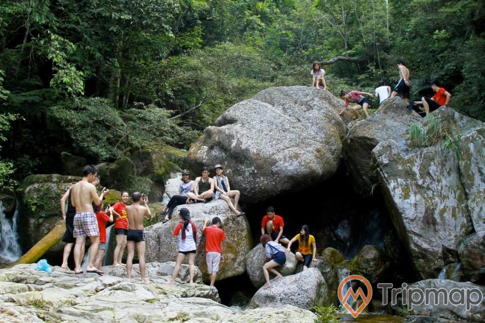 Nhóm du khách trải nghiệm tại thác Ba Tia, 1 nhóm người đang đứng chụp ảnh, 1 nhóm người khác ngồi trên phiến đá và 1 nhóm người đang đi trên phiến đá trên cao, ảnh chụp ngoài trời, cây cối phía sau phiến đá lớn
