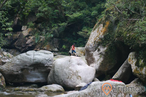 Vùng hạ lưu tại thác Ba Tia, phiến đá mọc rêu xanh, cây cối xanh tươi trên vách núi, 1 người phụ nữ áo đỏ đang đi bộ trên phiến đá, ảnh chụp ngoài trời