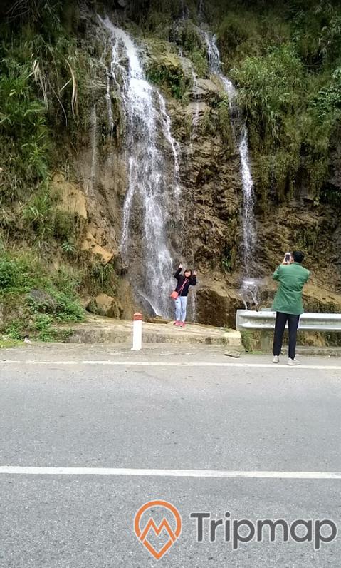 du khách chụp ảnh tại thác bạc, thác nước đang chảy, cây cối gần thác nước, chàng trai áo xanh lá đang đứng chụp cho cô gái đứng gần thác nước, ảnh chụp ngoài trời