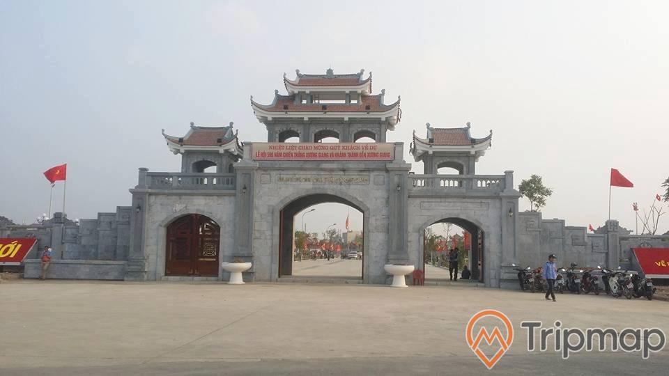 cổng thành khu di tích lịch sử chiến thắng Xương Giang, cổng thành bằng đá 2 tầng và có mái ngói màu đỏ, 2 lá cờ bay phất phới trên thành cổng, 2 chậu cây màu trắng ở phía trước cổng, nhiều xe máy và chú bảo vệ áo màu xanh ở phía trước cổng, bầu trời nhiều mây