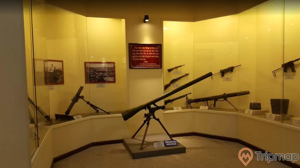 vũ khí thời chiến tranh trưng bày tại bảo tàng Chiến thắng Điện Biên Phủ, các loại súng treo trên tường và có 1 cây súng cối đặt phía dưới, bức tranh treo trên tường, ánh đèn màu vàng, ảnh chụp trong nhà bảo tàng