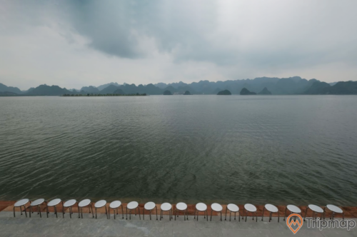 Quang cảnh yên bình tại khu du lịch hồ Tam Chúc, hàng ghế inox trên bậc thềm bờ hồ, dãy núi đồi phía xa xa, bầu trời nhiều mây đen, ảnh chụp ngoài trời