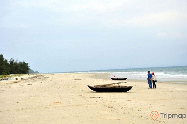Bãi biển Cồn Khởi, bãi cát trắng, chiếc thuyền gỗ trên bãi cát, hai người đang đứng trên bãi cát, ảnh chụp ban ngày