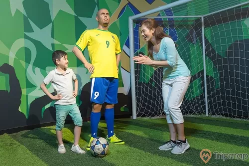 Bảo tàng tượng sáp, tượng sáp cầu thủ Ronaldo, em bé mặc áo trắng đang chống tay, quả bóng đá trên thảm cỏ màu xanh, người phụ nữ đang vỗ tay, khung thành bên cạnh