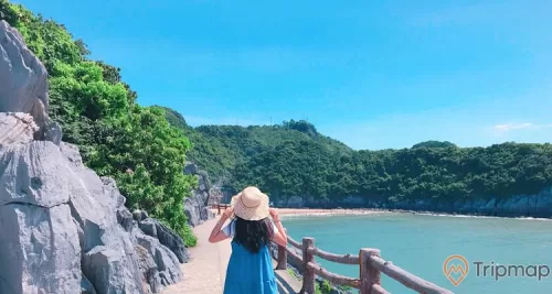 Đảo Cát Bà, cô gái mặc váy xanh đội mũ, tảng đá màu xám, lan can bằng gỗ, con đường màu xám, nhiều cây xanh, hồ nước, ngọn núi phía xa, ảnh chụp ban ngày