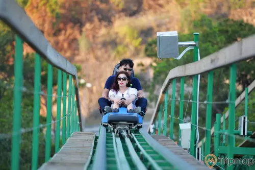 Khu làn trượt Samurai, Dragon Park, đường ray sơn màu xanh, 2 người đang ngồi trên ghế trượt, ngọn núi phía sau, trời nắng, ảnh chụp ban ngày