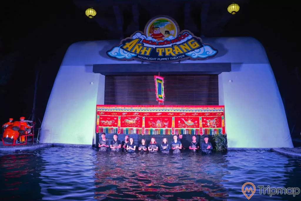 Sân khấu múa rối nước Ánh Trăng, nhiều người mặc áo đen đang đứng dưới nước, một vài người mặc đồ đỏ đang đánh trống, đèn lồng màu vàng, bức tường màu trắng